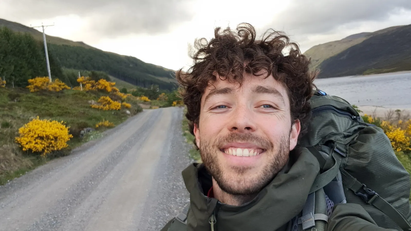 Cameron smiling on a gravel road in the hills, wearing a backpack